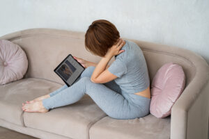 Woman sitting on her sofa and rubbing her painful neck while searching for cervical herniated disc symptoms on her tablet